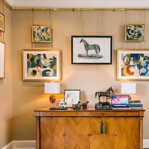 Könnte beinhalten: Innenaufnahme eines Zimmers mit einem Holzschrank, Lampen und Kunstwerken. An der Wand sind gerahmte Kunstwerke zu sehen, darunter abstrakte Gemälde und eine Pferdeillustration. Ein Laptop und Bücher stehen auf dem Schrank.