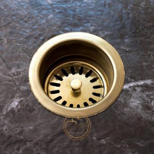 May include: A close-up of a brass sink drain with a circular design and a central knob. The drain has a series of small holes around the center. The background is a dark, marbled surface.