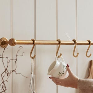 Puede incluir: Una barra de cocina dorada con varios ganchos en forma de S, que sostiene una jarra de cerámica blanca con un diseño floral verde. Cucharas medidoras cuelgan de un gancho. La barra está montada en una pared blanca panelada.