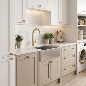 Puede incluir: Una cocina con armarios superiores blancos y armarios inferiores beige claro con pomos de latón. Un fregadero de acero inoxidable tipo granja se combina con un grifo alto de latón. El salpicadero tiene azulejos blancos en espiga e iluminación.
