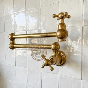 Puede incluir: Un grifo de latón para llenar ollas con un diseño vintage. El grifo presenta un brazo de doble articulación y manijas en forma de cruz. El grifo está montado en una pared de azulejos blancos.