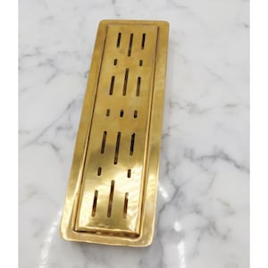 May include: A rectangular, brass-colored drain cover with a series of vertical and horizontal slots. The drain cover is set against a white and gray marble background. The brass finish gives the drain cover a vintage look.
