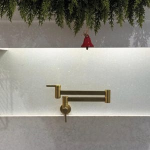 Peut inclure: Un robinet de remplissage de pot mural doré avec un bras articulé. Le robinet est installé sur une surface de couleur claire. Un petit ornement de cloche rouge est suspendu à une guirlande verte au-dessus.