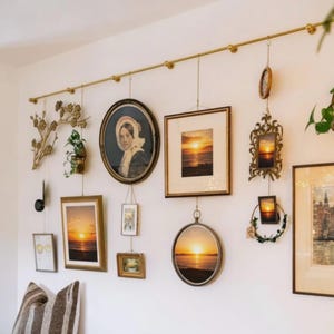 May include: A collection of framed art and decorative items hanging from a gold-colored rod on a white wall. The frames vary in shape and size, including oval and rectangular, and feature images of landscapes and portraits. Some frames are suspended by chains.