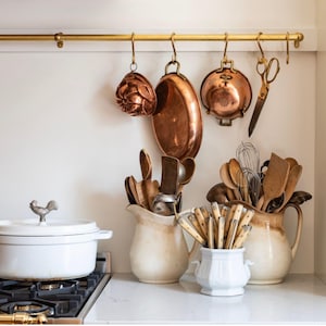 May include: A kitchen scene with copper cookware hanging from a gold rod. White ceramic pitchers hold wooden utensils and a white pot with a rooster-shaped handle sits on a stovetop. A pair of gold scissors hangs on the rod.