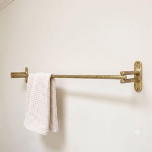 May include: A wall-mounted towel bar with a textured, brushed gold finish. The horizontal rod is supported by two side brackets that attach to the wall with visible screws. A folded, off-white waffle-weave towel hangs over the metal bar.