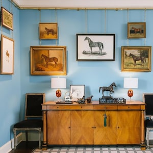 Pode incluir: Um quarto com paredes azul claro, adornado com pinturas de cavalos emolduradas e um armário de madeira. O armário é encimado por lâmpadas, esculturas e objetos decorativos. Duas cadeiras pretas estão posicionadas em frente ao armário.