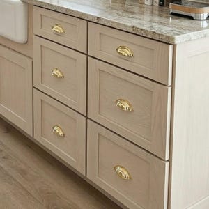 May include: Close-up of a light-coloured wooden kitchen cabinet with six drawers. Each drawer has a gold-coloured, half-moon shaped handle. The countertop is a speckled granite. The cabinet is part of a kitchen island.