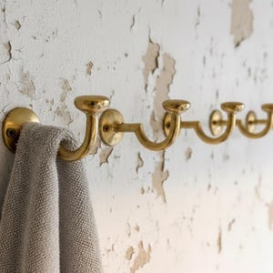 May include: A row of brass wall hooks with a towel hanging on one. The hooks have a curved design and are mounted on a distressed white wall. The towel is a light gray color.