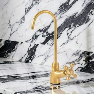 May include: A gold-colored faucet with a curved spout and a cross-shaped handle. The faucet is set against a backdrop of white and black marble. The fixture is a modern design element for a kitchen or bathroom.