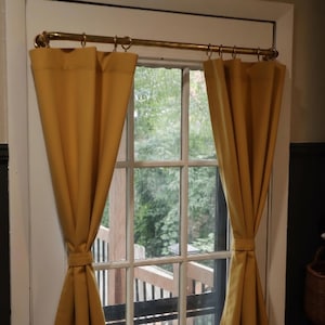 May include: Mustard yellow curtains hanging from a brass rod with rings. The curtains are tied back, revealing a window with a view of a wooden deck and lush greenery. The window frame is white.
