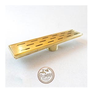 May include: A rectangular brass shower drain with multiple horizontal slots. The drain has a rounded base and is made of a shiny, gold-colored metal. The Plumbra logo is visible in the bottom right corner.