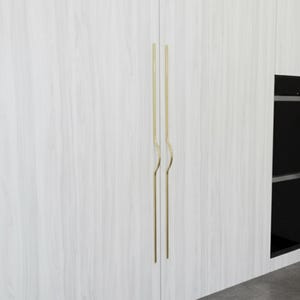 Może przedstawiać: Zbliżenie na jasną drewnianą szafkę z dwoma długimi, złotymi, zakrzywionymi uchwytami. Uchwyty są ustawione pionowo i mają smukły, elegancki design. Szafka jest częścią kuchni lub jednostki magazynowej.