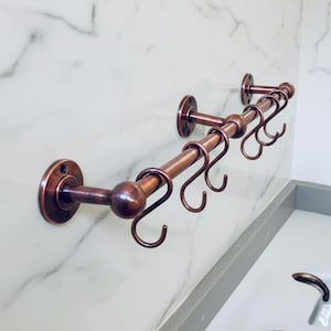 May include: A copper-colored metal towel rack with multiple curved hooks. The rack is mounted on a white marble wall. The design features a series of hooks for hanging towels or other items, adding a touch of elegance to any bathroom.
