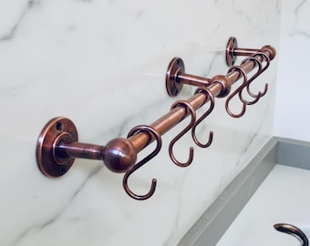 May include: A copper-colored metal towel rack with multiple curved hooks. The rack is mounted on a white marble wall. The design features a series of hooks for hanging towels or other items, adding a touch of elegance to any bathroom.