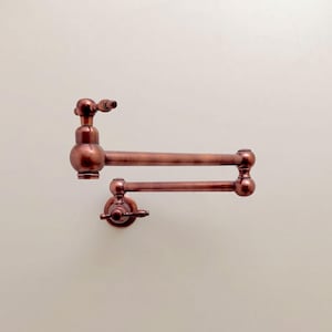 Könnte beinhalten: Ein wandmontierter Topffüller-Wasserhahn mit metallischem Kupfer-Finish. Die Armatur verfügt über ein doppeltes, gelenkiges Schwenkarm-Design mit zwei runden Griffen, einer an der Basis und einer am Auslauf, die durch zylindrische Rohre verbunden sind.