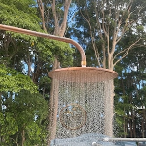 May include: Outdoor shower fixture with a copper-colored shower head and curved pipe. Water streams down from the circular shower head. The shower is set against a backdrop of green trees and a blue sky.