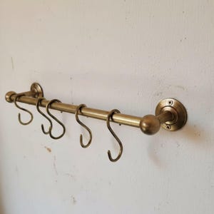 Może przedstawiać: Mosiężna szyna kuchenna z sześcioma haczykami w kształcie litery S. Szyna jest zamontowana na białej ścianie. Konstrukcja posiada zaokrąglony koniec i ozdobne okrągłe mocowania.