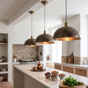 Peut inclure: Trois lampes suspendues de style antique avec des abat-jours en forme de dôme de couleur bronze sont suspendues au-dessus d'un îlot de cuisine. La cuisine comprend un comptoir en marbre, une crédence carrelée et une planche à découper en bois avec des herbes fraîches.