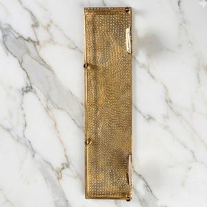 Macro close-up detail of solid brass showing the hand-hammered, dimpled texture. The unlacquered surface reflects light, highlighting the artisanal craftsmanship.