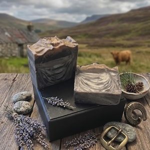May include: Two rectangular bars of marbled gray and white soap with textured tops, displayed on a black box alongside dried lavender sprigs, two smooth gray stones, a small ceramic bowl with pinecones, and a brass belt buckle.