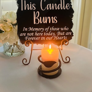 May include: A black sign with white text that reads "This Candle Burns In Memory of those who are not here today, But are Forever in our Hearts". A lit candle sits on a metal candle holder.