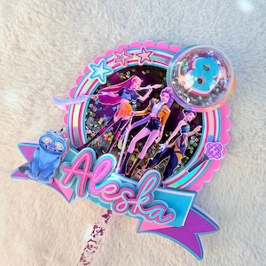 May include: A colorful cake topper with the name "Alaska" in pink and blue script. The topper features a cartoon image of three figures, a blue cat, and a clear bubble with the number 8 inside. The design includes pink, blue, and purple accents.