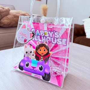 May include: Clear plastic tote bag with a handle, featuring a pink and white Gabby's Dollhouse design. The bag includes a purple car with Gabby and a cat character. A tag reads "Julietta's 5th Birthday."