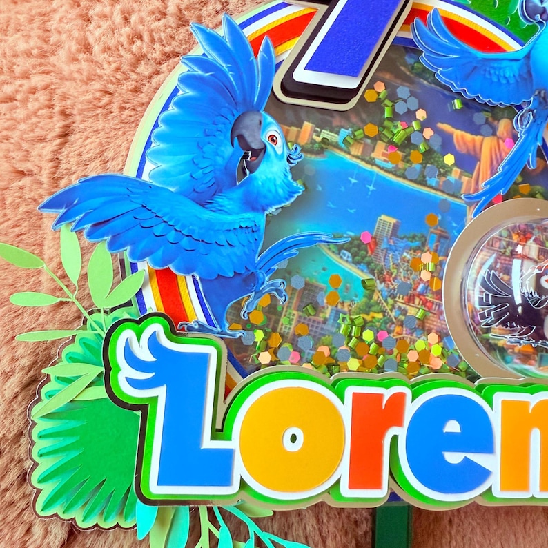 Op de afbeelding: Een kleurrijke taarttopper met een blauwe ara met uitgespreide vleugels. De topper heeft een Rio de Janeiro-thema, met de stad en de Christus de Verlosser zichtbaar. De naam "Loren" wordt in levendige kleuren weergegeven, met het cijfer "1" ervoor.