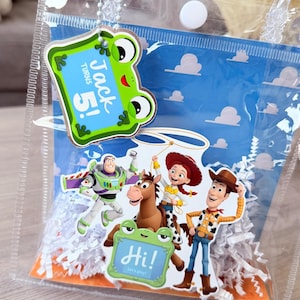 May include: A clear plastic gift bag with white paper shred filler, featuring a blue background with white clouds. Attached is a green frog tag reading Jack Turns 5! and a second tag reading Hi! Let's play! with Toy Story character cutouts.