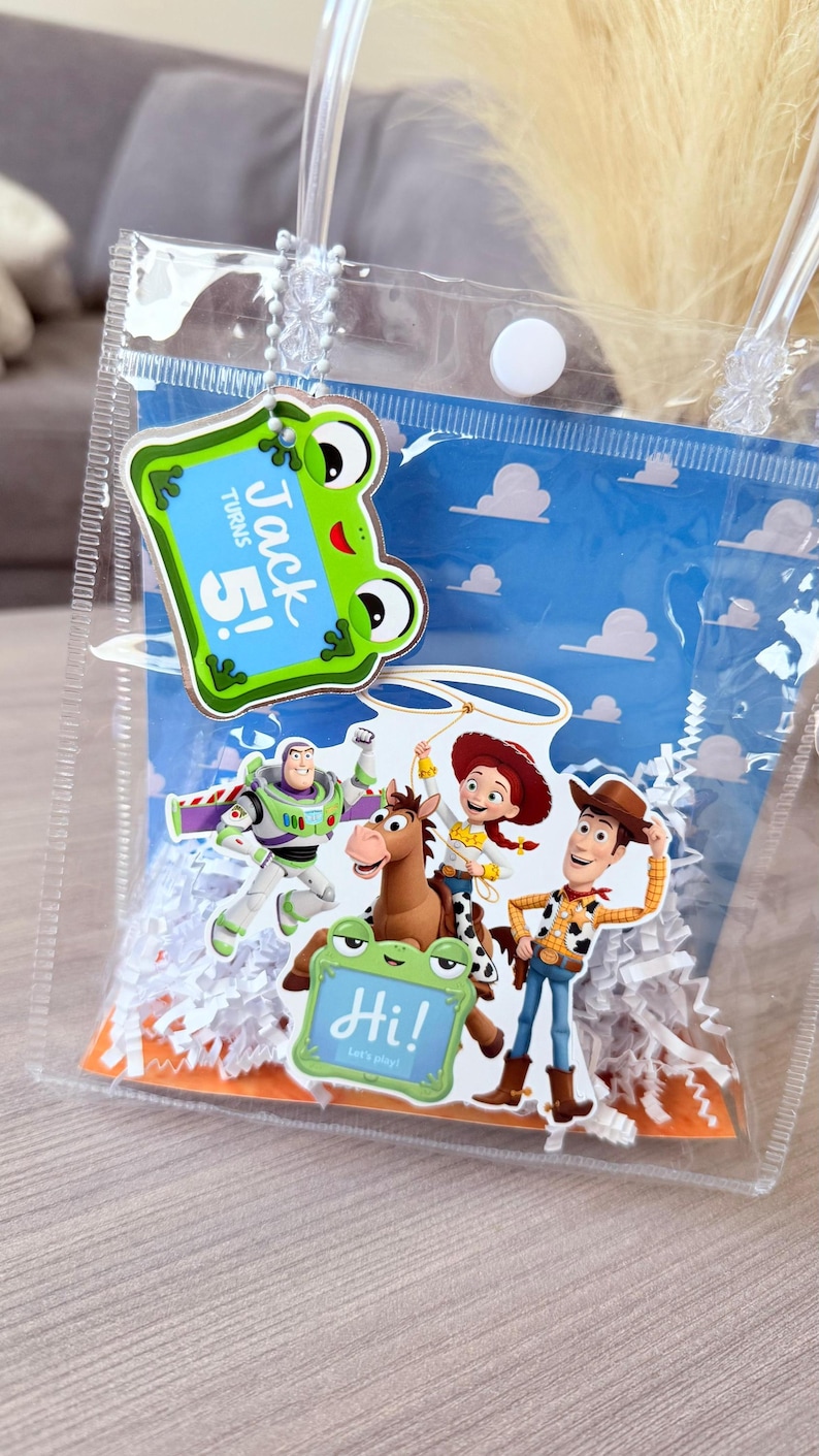 May include: A clear plastic gift bag with white paper shred filler, featuring a blue background with white clouds. Attached is a green frog tag reading Jack Turns 5! and a second tag reading Hi! Let's play! with Toy Story character cutouts.