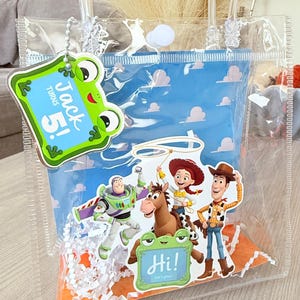 May include: A clear plastic gift bag with white handles, filled with white paper shred. It features a blue cloud-patterned insert, a green frog tag reading "Jack Turns 5!", and a cutout of Buzz, Woody, Jessie, and Bullseye with a "Hi!" tag.