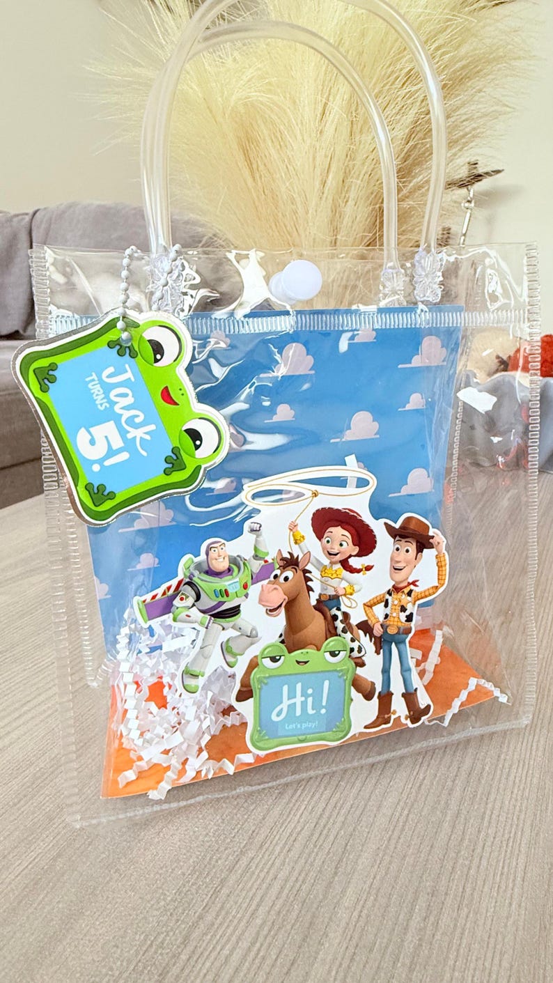 May include: A clear plastic gift bag with white handles, filled with white paper shred. It features a blue cloud-patterned insert, a green frog tag reading "Jack Turns 5!", and a cutout of Buzz, Woody, Jessie, and Bullseye with a "Hi!" tag.