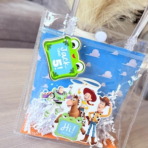 May include: A clear plastic gift bag with white paper shred filler, featuring a blue background with clouds. Attached are cutouts of Toy Story characters and a green frog-shaped tag with the text Jack turns 5! and a smaller tag saying Hi!