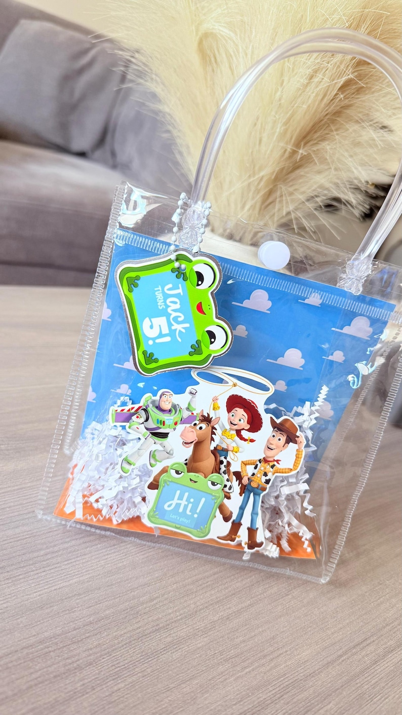 May include: A clear plastic gift bag with white paper shred filler, featuring a blue background with clouds. Attached are cutouts of Toy Story characters and a green frog-shaped tag with the text Jack turns 5! and a smaller tag saying Hi!
