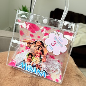 May include: Clear plastic tote bag with a handle, featuring a Moana theme. The bag has a pink floral pattern and a tag that says "Hannah's 3rd Birthday Party". The bag contains white decorative elements.