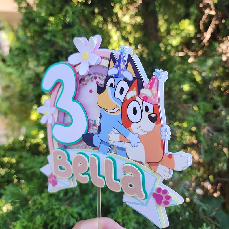 May include: A vibrant cake topper with the number 3, floral accents, and the name "Bella." It showcases Bluey and Bingo cartoon characters, with confetti visible. This topper is designed for a birthday celebration.