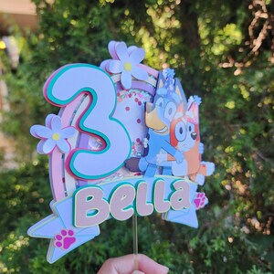 May include: A vibrant cake topper with the number "3", a rainbow, and flower details. The name "Bella" is displayed, along with cartoon dog characters. The design is predominantly pink, blue, and white, with a clear stem for placement.