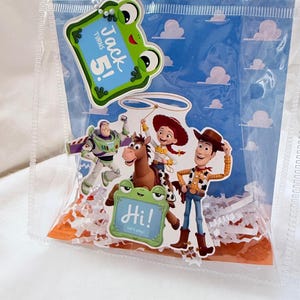 May include: A clear plastic gift bag with white handles and a snap closure. Inside is a blue background with white clouds, featuring Toy Story characters Buzz Lightyear, Jessie, and Woody. It includes two frog-shaped tags reading Jack turns 5! and Hi! Let's play!