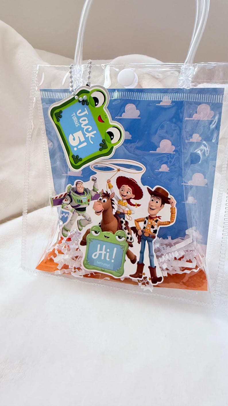 May include: A clear plastic gift bag with white handles and a snap closure. Inside is a blue background with white clouds, featuring Toy Story characters Buzz Lightyear, Jessie, and Woody. It includes two frog-shaped tags reading Jack turns 5! and Hi! Let's play!