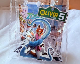 May include: A clear plastic gift bag featuring Zootopia characters. The bag includes a Zootopia-themed image with Nick Wilde and Judy Hopps. A tag reads "Olivia Turns 5". The bag is filled with white paper shreds, perfect for a birthday celebration.