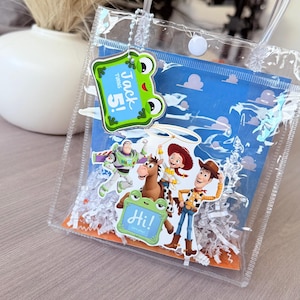 May include: A clear plastic gift bag with two handles and a white snap closure. Inside, a blue card features Toy Story characters including Buzz Lightyear, Woody, Jessie, and Bullseye. A green frog tag reads "Jack turns 5!" and "Hi! Let's play!"