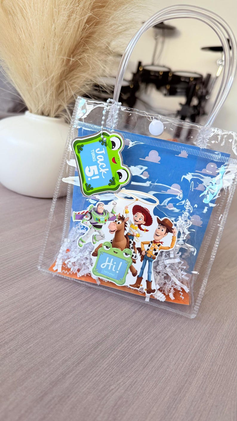 May include: A clear plastic gift bag with two handles and a white snap closure. Inside, a blue card features Toy Story characters including Buzz Lightyear, Woody, Jessie, and Bullseye. A green frog tag reads "Jack turns 5!" and "Hi! Let's play!"