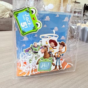 May include: A clear plastic gift bag with white handles and a snap closure. The bag features a blue background with a cloud pattern and cutouts of Toy Story characters including Buzz Lightyear, Jessie, Bullseye, and Woody. A green frog tag says Jack Turns 5!