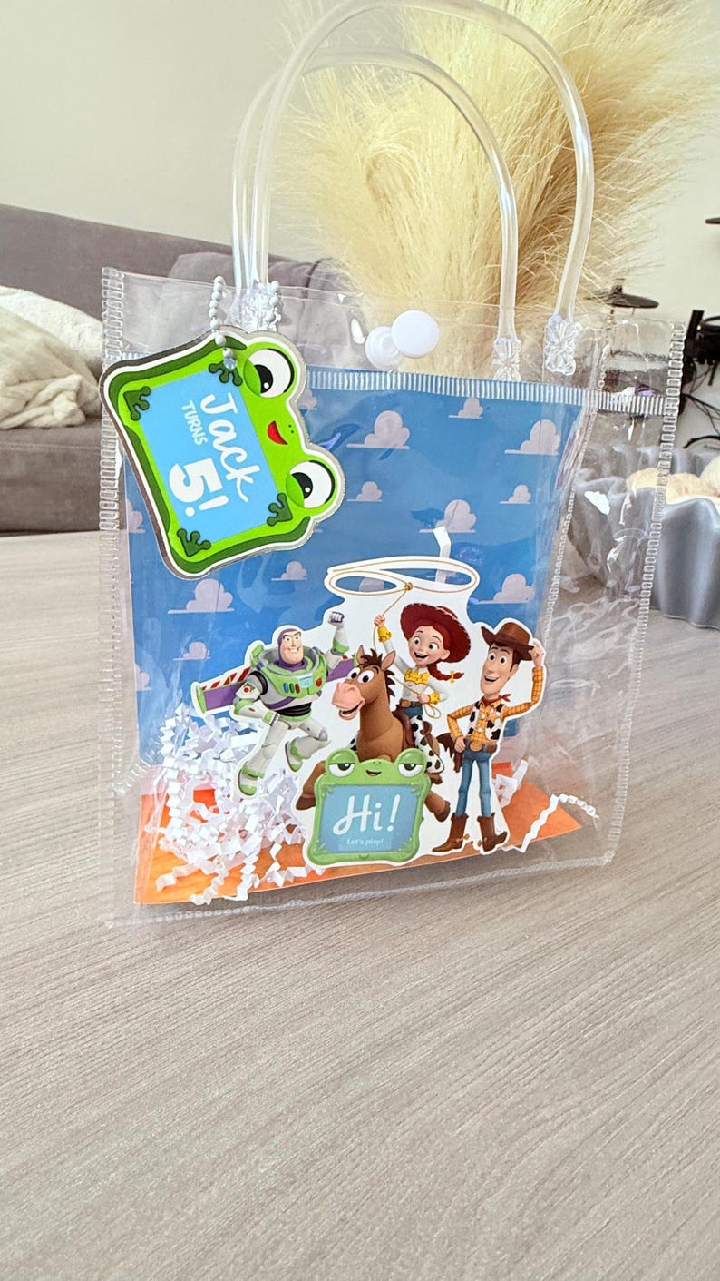 May include: A clear plastic gift bag with white handles and a snap closure. The bag features a blue background with a cloud pattern and cutouts of Toy Story characters including Buzz Lightyear, Jessie, Bullseye, and Woody. A green frog tag says Jack Turns 5!