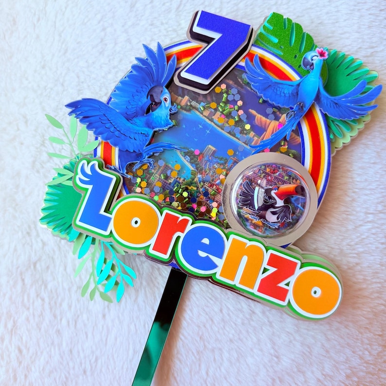 Op de afbeelding: Een kleurrijke taarttopper met de naam "Lorenzo" in oranje en blauw, het cijfer 7 en afbeeldingen van blauwe vogels. Het tropische thema bevat een regenboog en een toekan-illustratie, geschikt voor een verjaardag.