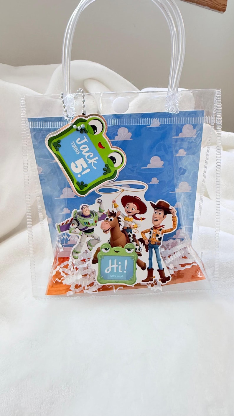 May include: A clear plastic gift bag with white handles and a snap closure. Inside, a blue sky and cloud background features Toy Story characters Buzz Lightyear, Jessie, Bullseye, and Woody. It includes a green frog tag reading Jack Turns 5! and a smaller green frog tag reading Hi! Let's play!