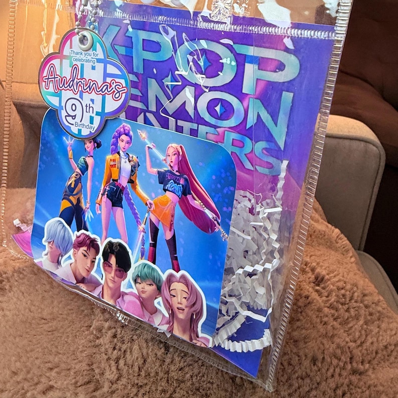 Puede incluir: Una bolsa transparente de celof&aacute;n llena de papel triturado blanco y una tarjeta decorativa. La tarjeta presenta un gr&aacute;fico colorido de figuras animadas y el texto "POP MONSTERS" y "9&ordm; Cumplea&ntilde;os de Audrina".