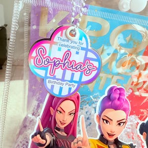 May include: A clear plastic bag with a cloud-shaped tag that reads "Thank you for celebrating Sophia's Birthday Party." The bag contains a colorful graphic of two animated women. The tag is attached to a white beaded chain.