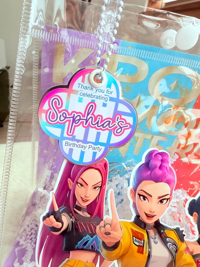 May include: A clear plastic bag with a cloud-shaped tag that reads "Thank you for celebrating Sophia's Birthday Party." The bag contains a colorful graphic of two animated women. The tag is attached to a white beaded chain.